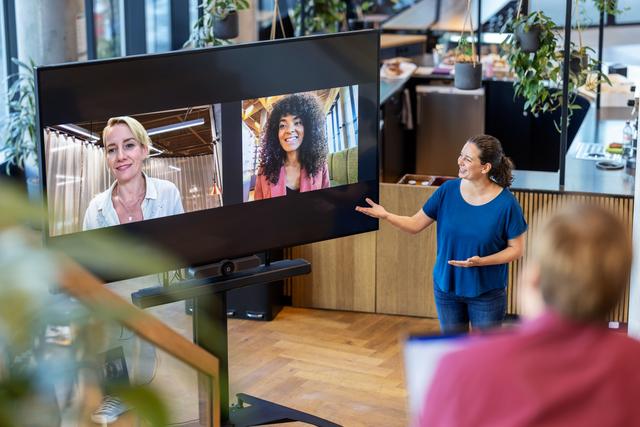 Smiling woman facilitates a hybrid event with two female guest speakers displayed on a big screen